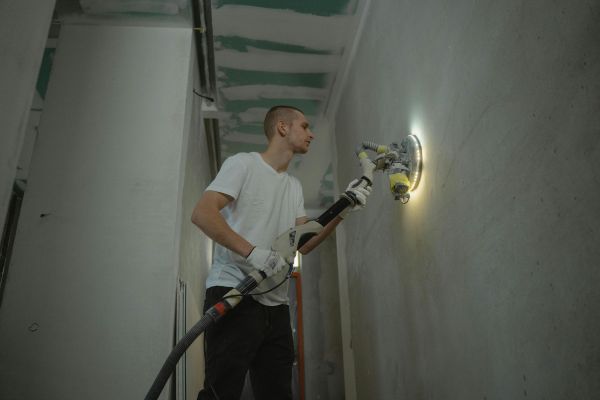 A man sanding an interior wall with an automatic sanding tool during renovation work indoors.